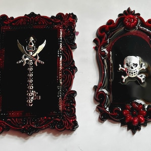 May include: Two decorative wall plaques with a gothic theme. One is rectangular with a skull and crossed swords key design. The other is oval with a skull and crossbones. Both have ornate red and black frames.