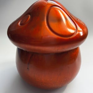 Puede incluir: Objeto decorativo brillante en forma de hongo, en tonos marrón rojizo. La parte superior tiene una tapa redondeada con hendiduras ovaladas, y la base es bulbosa. La superficie refleja la luz, dándole un aspecto pulido.