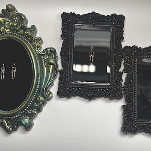 May include: Three ornate picture frames. An oval frame with a green and gold finish holds two small figures. Two rectangular black frames with intricate detailing each hold a single figure, creating a unique display.