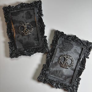 May include: Two ornate black picture frames with a textured, dark gray surface. Each frame features a silver-colored, stylized skull emblem in the center. The frames have detailed, decorative edges.