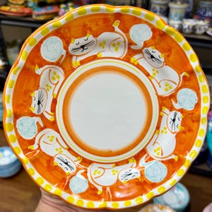 May include: A decorative ceramic plate with a vibrant orange background and a white center. The plate features playful white cat illustrations with yellow accents and a yellow dotted border. The plate's rim has a scalloped edge.