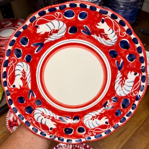 May include: A red ceramic plate with a white center and a scalloped edge. The plate is decorated with blue and white shrimp and blue dots. The plate is part of a set of dishes.