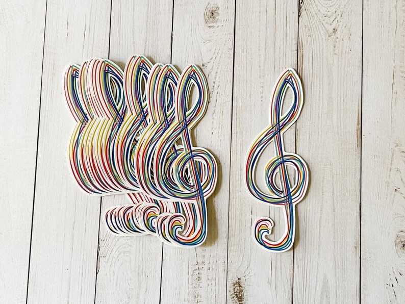 May include: A collection of colorful treble clef stickers. Each sticker features a white outline with a design of rainbow-colored lines. The stickers are arranged on a light-colored wooden surface.