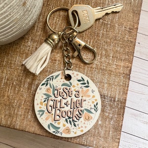 May include: A round wooden keychain with a floral design and the text "Just a Girl + her Books". The keychain has a gold-colored tassel and a metal key ring.