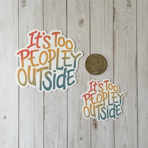 May include: Two stickers with the phrase "It's Too Peopley Outside" in a colourful, handwritten style. The larger sticker is approximately 7.6 cm wide, and the smaller one is about 5 cm wide. A coin is next to the stickers for size comparison.