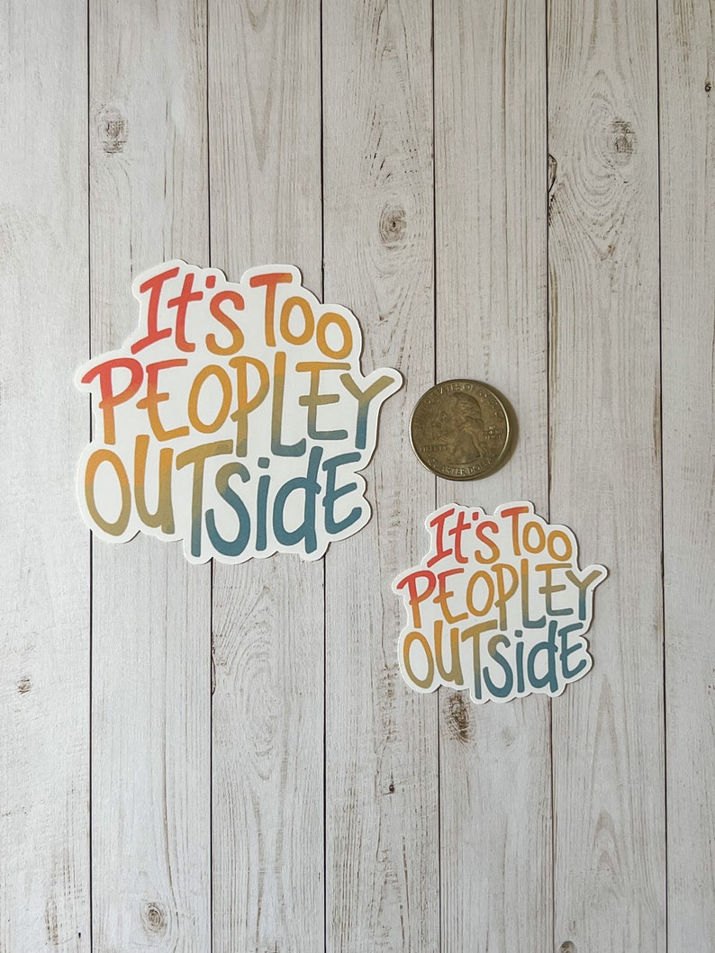 May include: Two stickers with the phrase "It's Too Peopley Outside" in a colourful, handwritten style. The larger sticker is approximately 7.6 cm wide, and the smaller one is about 5 cm wide. A coin is next to the stickers for size comparison.