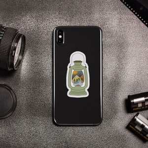 May include: A black smartphone with a sticker of a green lantern with a mountain scene inside. The lantern has a yellow handle and a yellow flame at the top. The phone is on a gray surface with a camera, film canisters, and a roll of film.