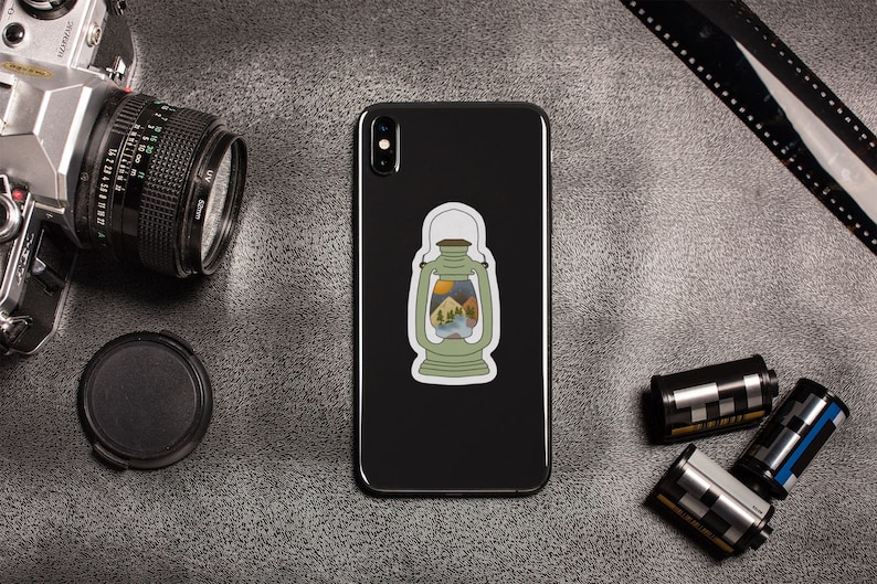 May include: A black smartphone with a sticker of a green lantern with a mountain scene inside. The lantern has a yellow handle and a yellow flame at the top. The phone is on a gray surface with a camera, film canisters, and a roll of film.
