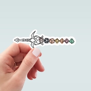 May include: A sticker featuring a black and white sword design with colorful dice along the blade. The dice are various shapes and colors, including gray, black, tan, yellow, pink, purple, and green. The sticker is held by a hand.