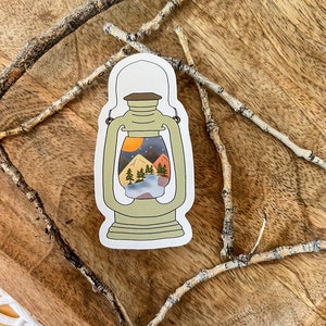 May include: A sticker of a green lantern with a scene of a mountain range, trees, and a moon inside. The sticker is on a wooden surface.
