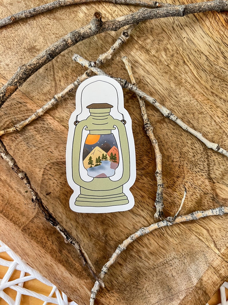 May include: A sticker of a green lantern with a scene of a mountain range, trees, and a moon inside. The sticker is on a wooden surface.