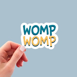May include: A white sticker with the word "WOMP" in teal and yellow, with a blue outline. The sticker is held by a hand with a light blue background.