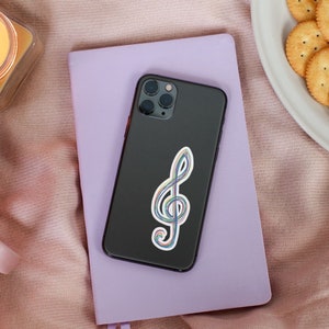 May include: A black smartphone with a colorful treble clef sticker rests on a lavender notebook. A lit candle in a glass jar, a pink mug with tea, and a plate of crackers are also in the image, set on a pink blanket.