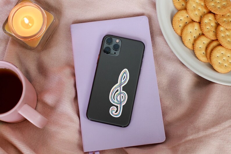 May include: A black smartphone with a colorful treble clef sticker rests on a lavender notebook. A lit candle in a glass jar, a pink mug with tea, and a plate of crackers are also in the image, set on a pink blanket.