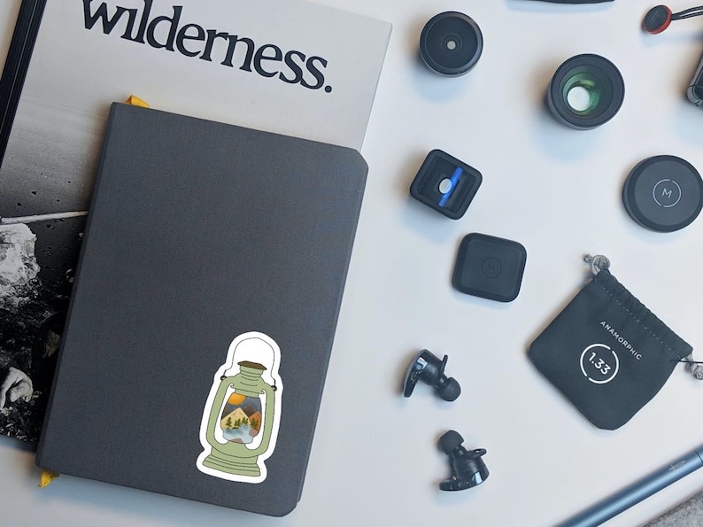 May include: A gray notebook with a sticker of a lantern with a mountain scene inside. The lantern is white with a brown handle and a brown base. The mountain scene is a colorful landscape with green trees, blue water, and brown mountains.
