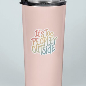 May include: A pale pink insulated travel mug with a black lid. The mug features the phrase "It's Too PEOPLEY OUTSIDE" in colourful, handwritten lettering. The mug is designed to keep drinks hot or cold, perfect for travel.