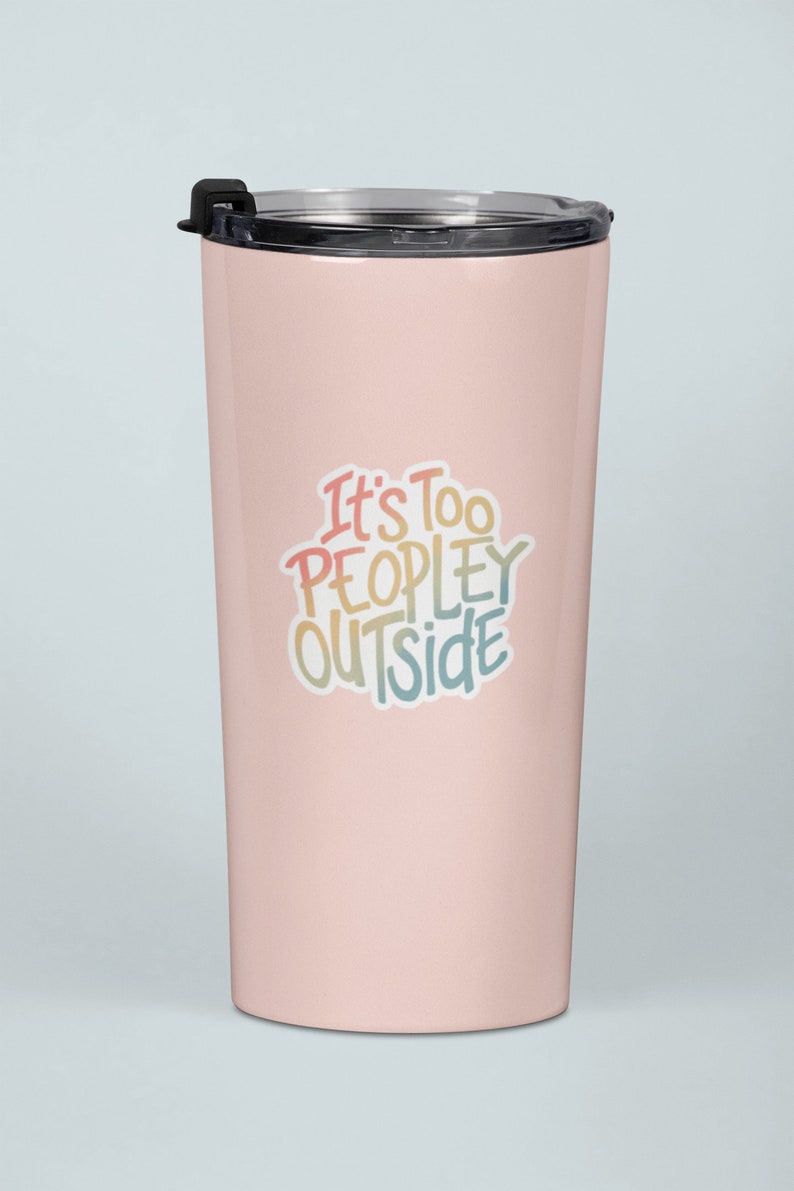 May include: A pale pink insulated travel mug with a black lid. The mug features the phrase "It's Too PEOPLEY OUTSIDE" in colourful, handwritten lettering. The mug is designed to keep drinks hot or cold, perfect for travel.