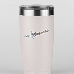 May include: A light pink insulated travel mug with a silver lid. A black and white line drawing of a sword with dice attached to the blade is on the front. The dice are various colours and shapes.