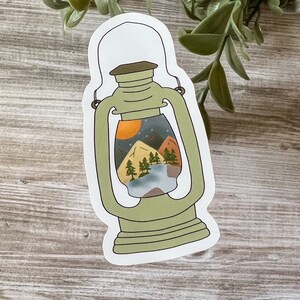 May include: A green lantern sticker with a mountain scene inside. The scene includes a lake, trees, and a sunset.