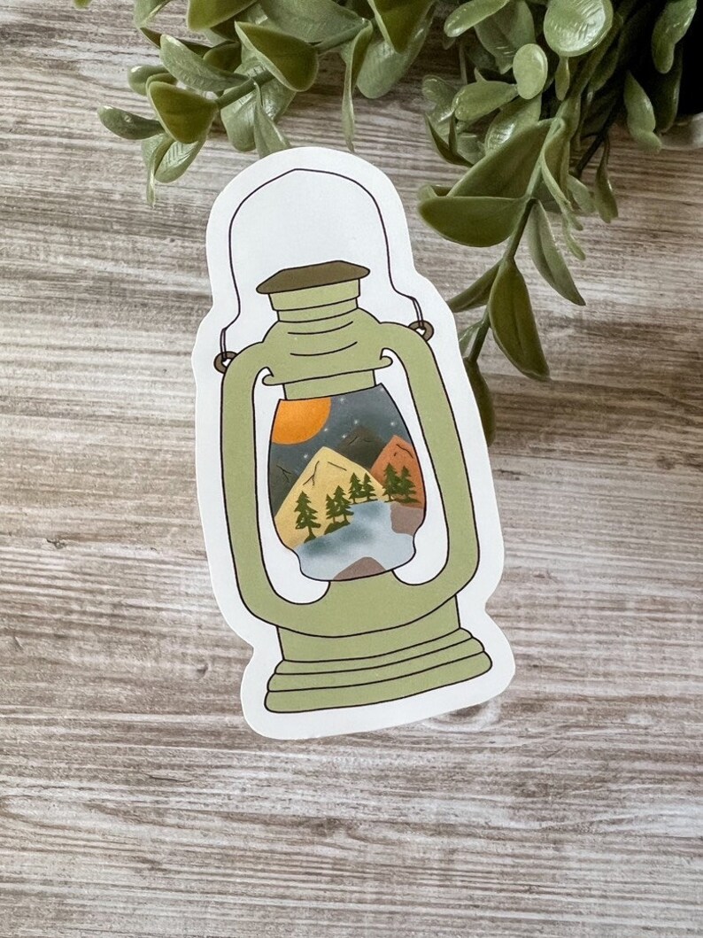 May include: A green lantern sticker with a mountain scene inside. The scene includes a lake, trees, and a sunset.