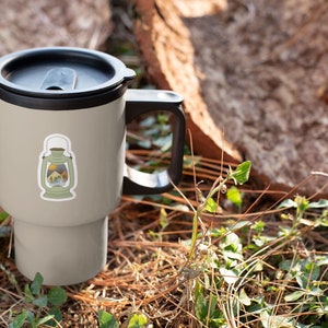 May include: A gray travel mug with a black lid and handle. The mug has a sticker of a lantern with a mountain scene inside. The mug is sitting on a bed of pine needles and green plants.