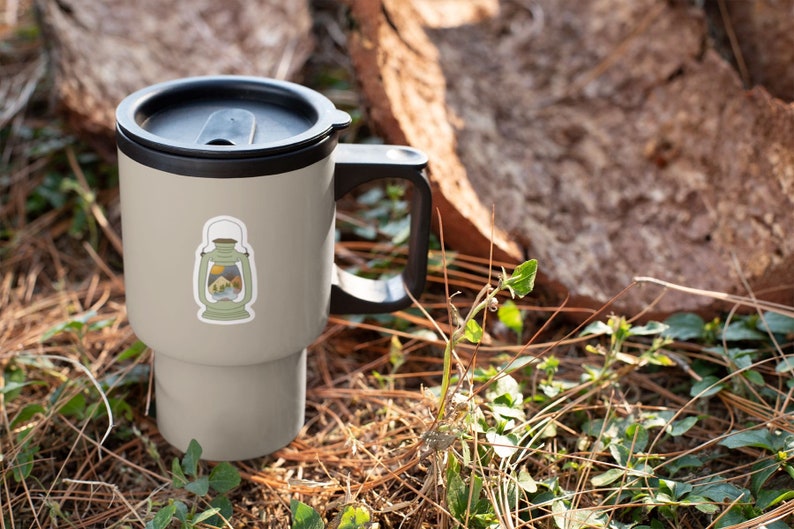 May include: A gray travel mug with a black lid and handle. The mug has a sticker of a lantern with a mountain scene inside. The mug is sitting on a bed of pine needles and green plants.