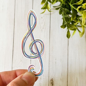 May include: A colorful treble clef sticker with a rainbow design. The musical symbol is outlined with thin lines in red, blue, yellow, and green. The sticker is held against a white wooden background with green foliage.