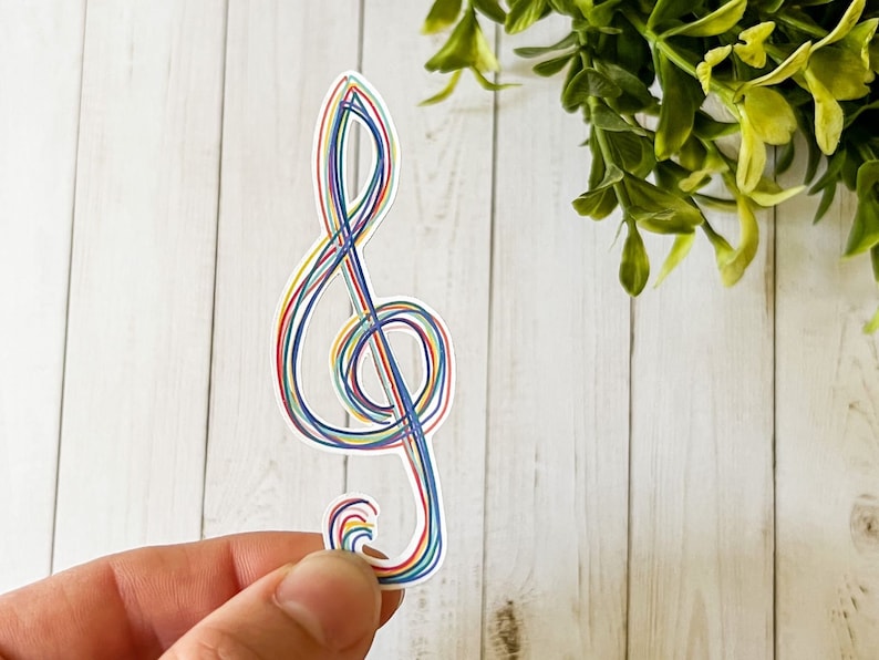 May include: A colorful treble clef sticker with a rainbow design. The musical symbol is outlined with thin lines in red, blue, yellow, and green. The sticker is held against a white wooden background with green foliage.