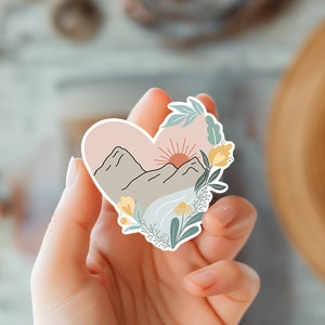 May include: A heart-shaped sticker with a mountain landscape, a sun setting behind the mountains, and a river flowing through the valley. The sticker is surrounded by green leaves and yellow flowers.