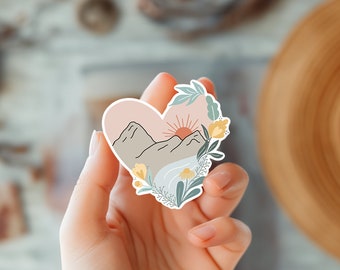 May include: A heart-shaped sticker with a mountain landscape, a sun setting behind the mountains, and a river flowing through the valley. The sticker is surrounded by green leaves and yellow flowers.