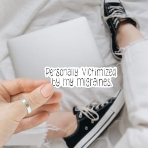 May include: A white sticker with black text that reads "Personally Victimized by my Migraines." The sticker is being held by a person's hand wearing a silver ring.