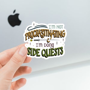 May include: A sticker with a whimsical design featuring the text "I'm not procrastinating, I'm doing side quests." The text is in a stylized font with gold and green accents. The sticker is on a white background.