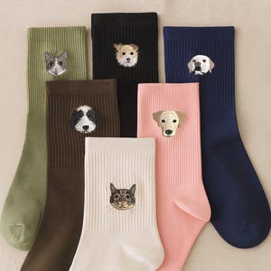 Puede incluir: Una colección de calcetines coloridos, cada uno con una cara de animal bordada. Los calcetines son de varios colores, incluyendo verde oliva, negro, marrón, blanco, rosa y azul marino. Los animales bordados incluyen gatos y perros.