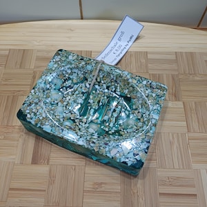 Può includere: Un portasapone quadrato con un design a mosaico nei toni del verde acqua, beige e bianco. Il portasapone ha un portasapone integrato ed è realizzato in un materiale trasparente e strutturato. L'etichetta recita "Seifenschale groß".