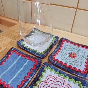 Könnte beinhalten: Vier handgefertigte Stoffuntersetzer mit einem blauen und grünen karierten Rand. Jeder Untersetzer zeigt ein anderes gesticktes Design in rotem und grünem Garn. Die Untersetzer liegen auf einer Holzoberfläche mit einem Glas auf einem der Untersetzer.