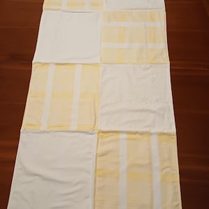 Könnte beinhalten: Ein Patchwork-Tischläufer mit abwechselnden Quadraten aus weißem und gelbem Stoff. Einige Quadrate haben ein gelb-weiß gestreiftes Muster, während andere einfarbig weiß sind oder gestickte Blumenmuster aufweisen. Der Läufer liegt auf einer dunklen Holzoberfläche.