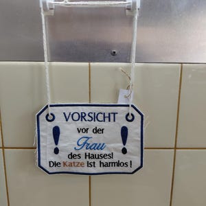 Könnte beinhalten: Ein rechteckiges weißes Stoffschild mit blauen Stickrändern und dem Text: VORSICHT vor der Frau des Hauses! Die Katze ist harmlos! Das Schild zeigt zwei große blaue Ausrufezeichen und hängt an einer weißen Schnur, die an zwei Haken befestigt ist.