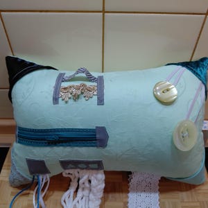 Puede incluir: Un cojín sensorial rectangular de color verde claro con varios elementos táctiles, incluyendo una cremallera azul cerceta, dos botones grandes translúcidos, un borde de encaje decorativo, un asa de tela pequeña y hebras de lana blanca.