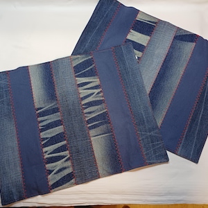 Könnte beinhalten: Zwei quadratische Kissenbezüge aus Denim. Jeder Kissenbezug besteht aus Streifen aus blauem Denim-Stoff, die mit rotem Faden zusammengenäht sind. Das Design umfasst unifarbene blaue Stoffstreifen. Die Kissenbezüge liegen auf einer weißen Oberfläche.