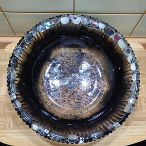 Könnte beinhalten: Eine dekorative Glasschale mit dunklem, rauchigem Aussehen. Die Schale hat einen strukturierten Rand und ist mit kleinen, klaren, irisierenden Perlen verziert. Der Innenraum weist ein gesprenkeltes Design mit Silber- und Blaunuancen auf.