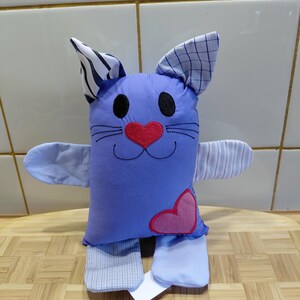 Può includere: Un gatto di peluche realizzato in tessuto azzurro con un naso a forma di cuore ricamato in rosso e una toppa a forma di cuore rosa. Le orecchie presentano motivi a righe bianche e nere e a quadretti blu, mentre braccia e gambe hanno righe.