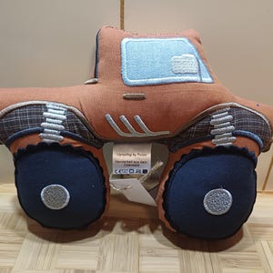 May include: A plush, brown toy truck with blue and gray accents. The truck features large, dark blue wheels with silver embroidered details. The cab has a light blue window, and the design includes plaid fabric details. A tag is attached.