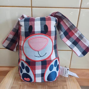 Könnte beinhalten: Ein Türstopper in Form eines Hasen aus Stoff mit einem rot-weiß-marineblauen Karomuster. Der Hase hat ein hellblaues Gesicht mit einer rosa Nase und gestickten Details. Die Pfoten sind marineblau mit rosa Akzenten, und die Ohren sind lang und schlaff.