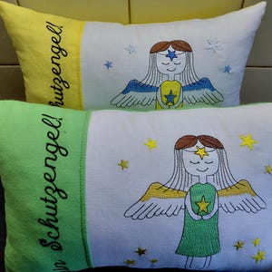 May include: Two rectangular throw pillows with white centers and colored side panels. The top pillow has a yellow side panel and the bottom has a green side panel. Both feature embroidered angel figures, stars, and the text "Schutzengel!" in black.