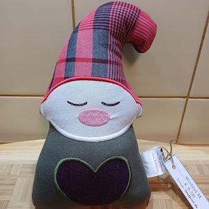Könnte beinhalten: Eine skurrile Wichtelpuppe mit kariertem rot-blauem Hut, weißem Gesicht, geschlossenen Augen und einer rosa Nase. Die Puppe hat einen grünen Körper mit einem herzförmigen Applikation in Lila. Ein Etikett mit Text ist angebracht.