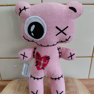 Könnte beinhalten: Eine rosa Stoffpuppe mit einem aufgenähten Design. Die Puppe hat ein großes, einzelnes Auge, aufgenähte Kreuzmarkierungen und einen herzförmigen Aufnäher. Die Puppe besteht aus rosa gestreiftem Stoff mit weißen Punkten. Ein Etikett ist angebracht.