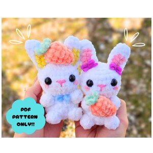 May include: Two white crocheted bunny plushies with colorful yarn details. One bunny is holding a pink and orange crocheted carrot. The other bunny has a pink, yellow, and orange crocheted flower on its head. The bunnies are sitting in a person's hands.