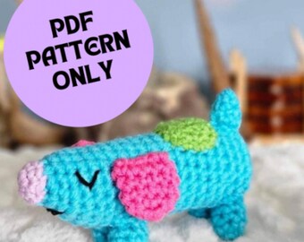 May include: A crocheted amigurumi dachshund toy with a blue body, pink ears, and green spots. The toy is lying on a white fluffy surface. The text "PDF PATTERN ONLY" is visible in the background.
