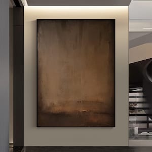 May include: Abstract art piece with a dark brown and tan color palette. The artwork is rectangular and framed in black. The painting has a textured appearance with a gradient effect, transitioning from lighter tones at the top to darker shades at the bottom.