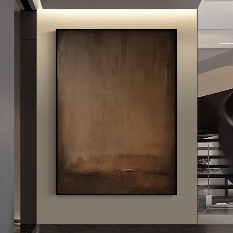 May include: Abstract art piece with a dark brown and tan color palette. The artwork is rectangular and framed in black. The painting has a textured appearance with a gradient effect, transitioning from lighter tones at the top to darker shades at the bottom.